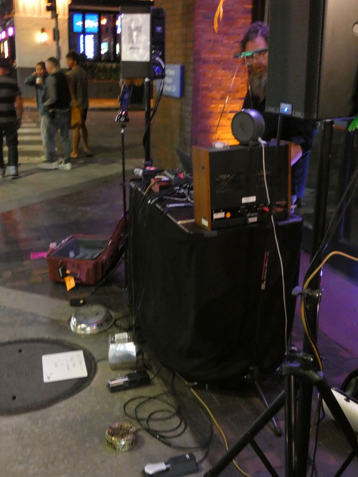 Side view of the rig on a sidewalk at night, a reel-to-reel deck on a draped table, cables and an open case on the ground, people passing on the street