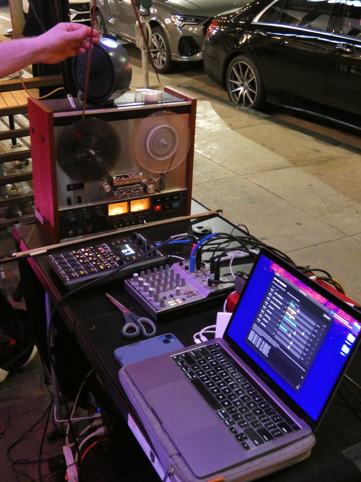 A hand threading tape onto a reel-to-reel deck on a gear table beside a sampler, mixer and laptop, cars parked along the street