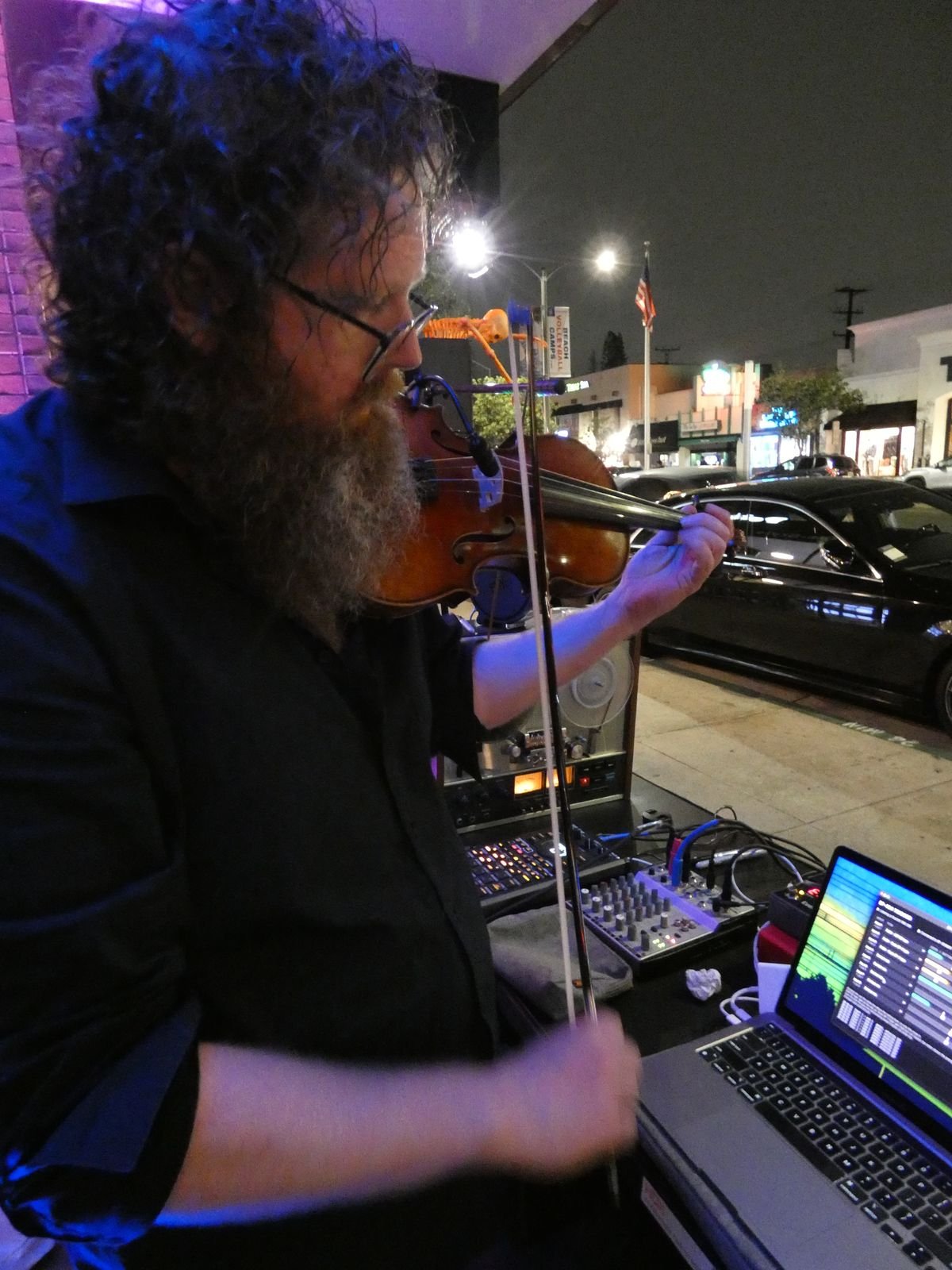 Aaron playing violin at night on Second Street in Long Beach, with a reel-to-reel deck, sampler, mixer and laptop in front of him and street lights behind