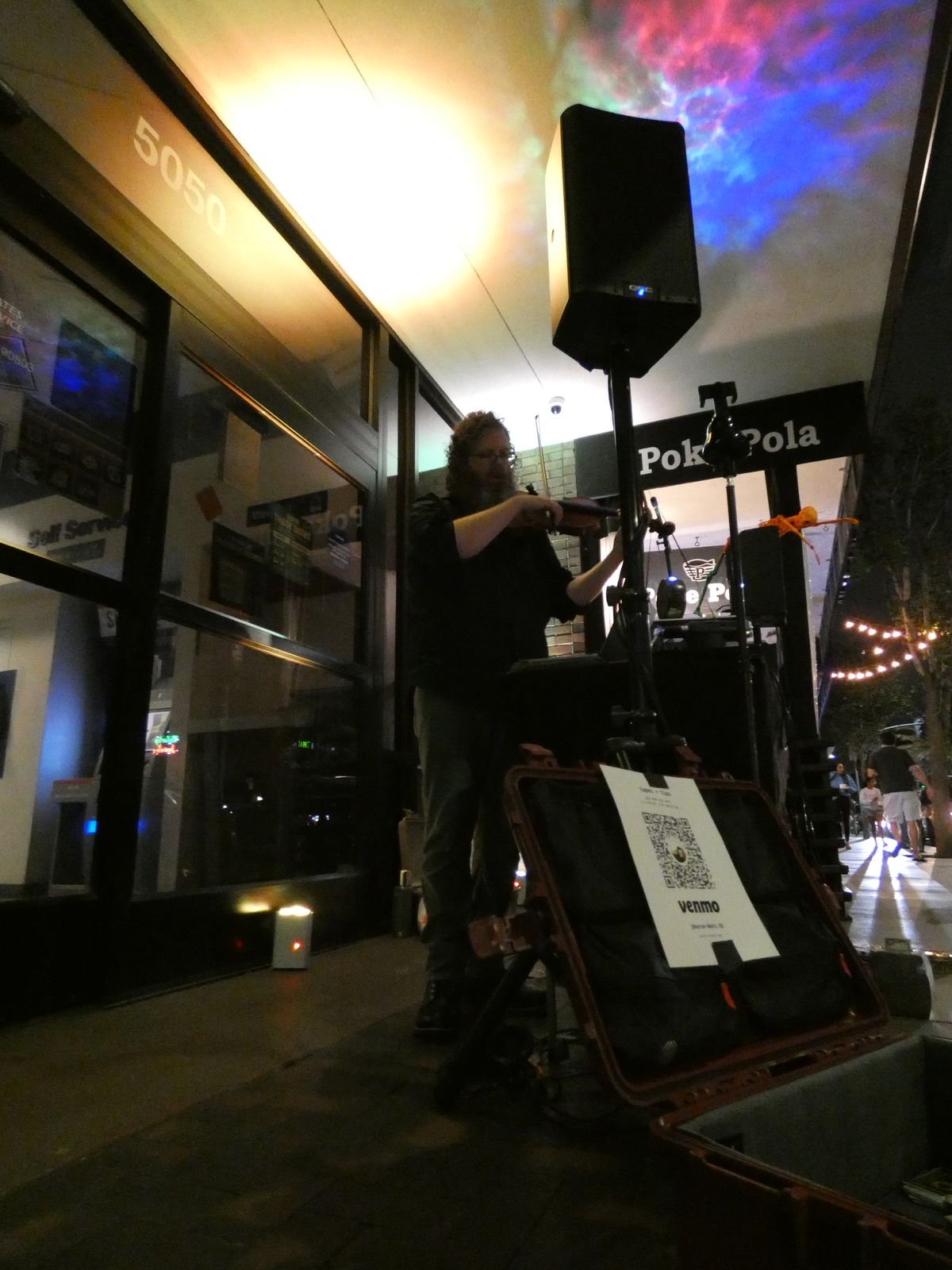 Low wide view of Aaron playing violin under a tall speaker on a sidewalk at night, colored light projected across the ceiling above