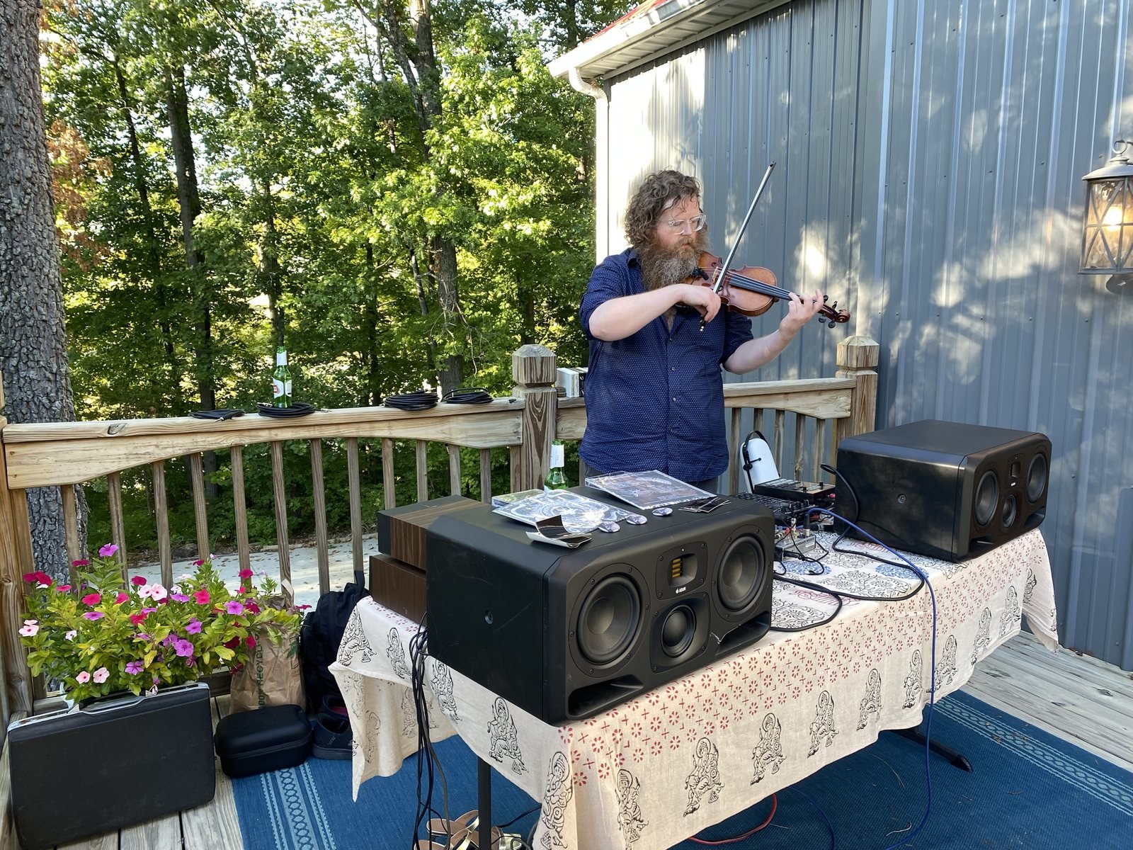 Aaron playing violin at a table draped in block-printed cloth, with large studio monitors, cassettes and small pins on top, on a wooden deck among trees