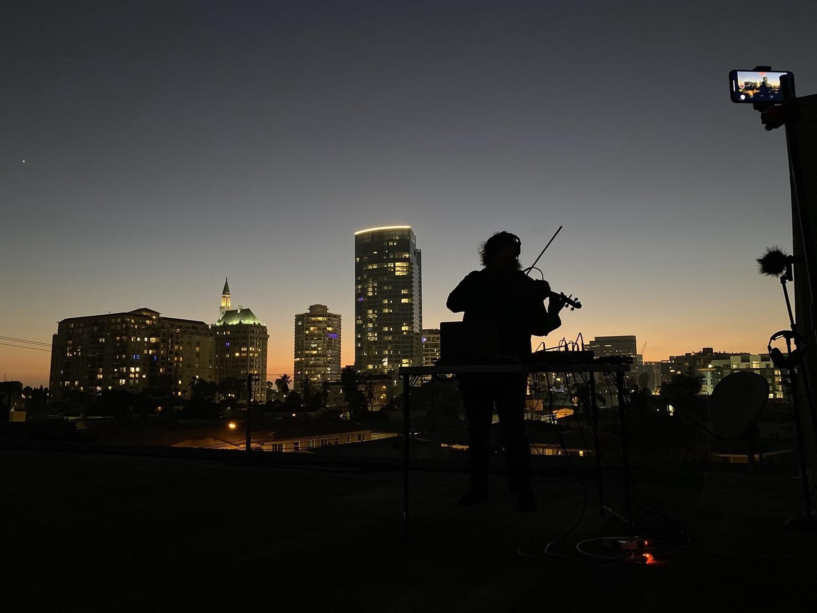 Silhouette of Aaron Nicholas West playing violin at a table of equipment on a rooftop at dusk, with a city skyline behind