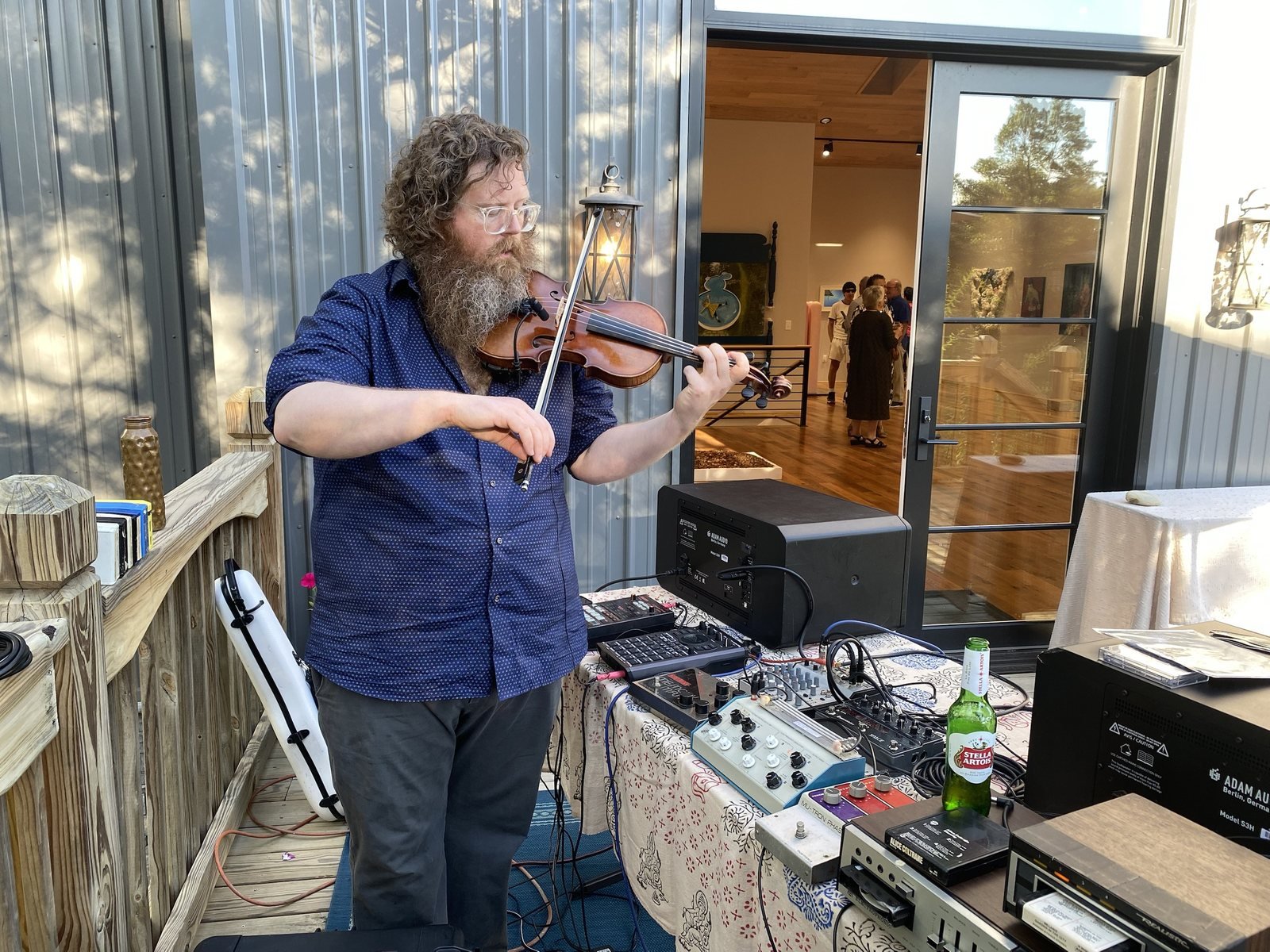 Aaron Nicholas West playing violin beside a table of samplers, pedals and 8-track players on a wooden deck, with gallery guests visible through an open door behind him