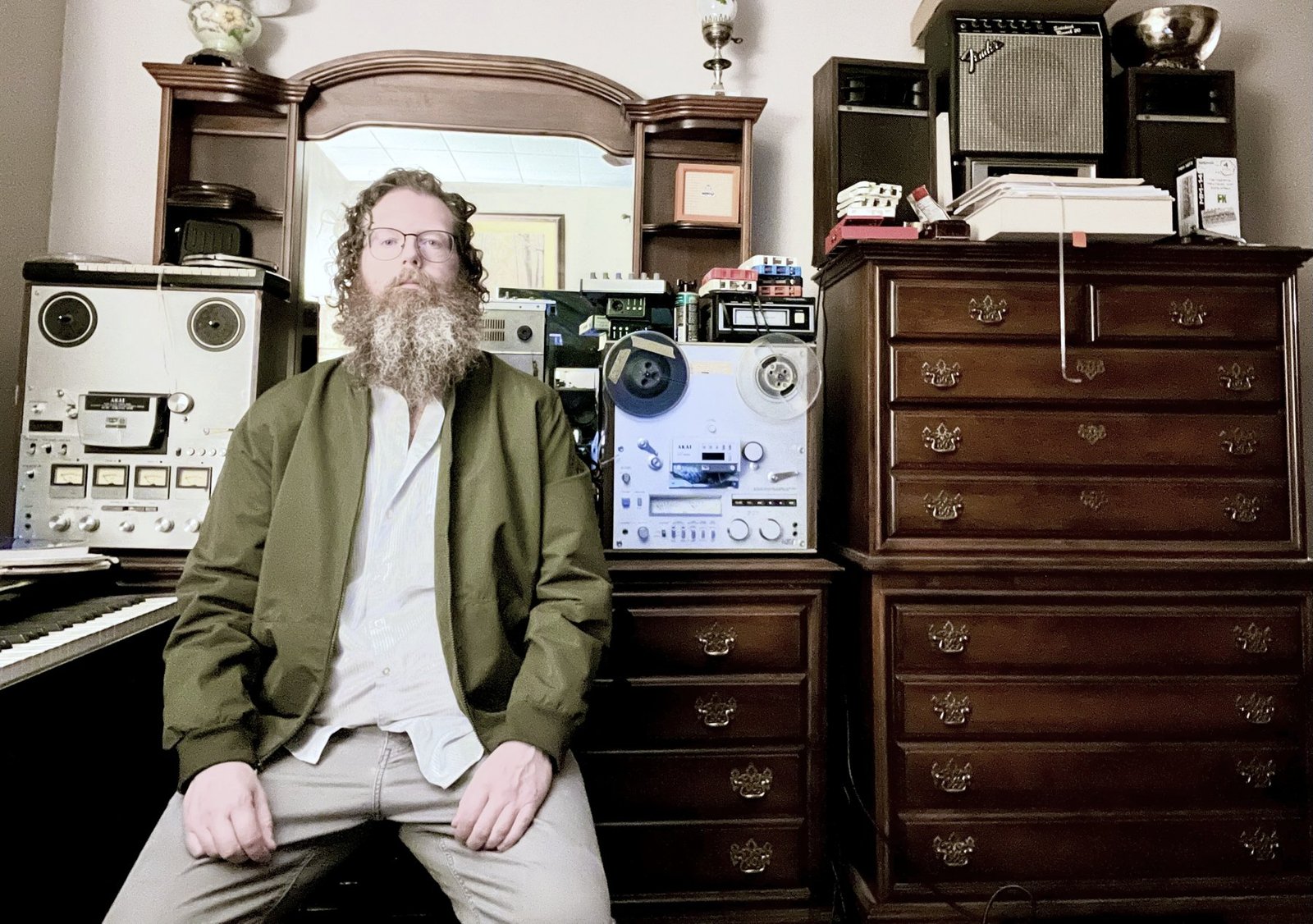 Aaron Nicholas West seated in his studio in a green bomber jacket, between two reel-to-reel tape machines and a tall wooden chest of drawers