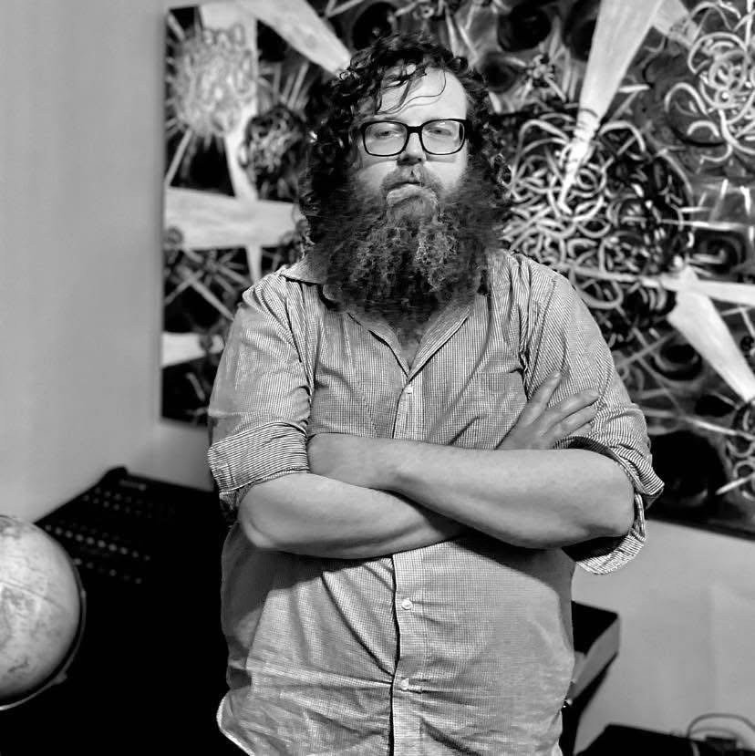 Black and white portrait of Aaron Nicholas West with arms crossed in front of a dark abstract painting, a globe at lower left