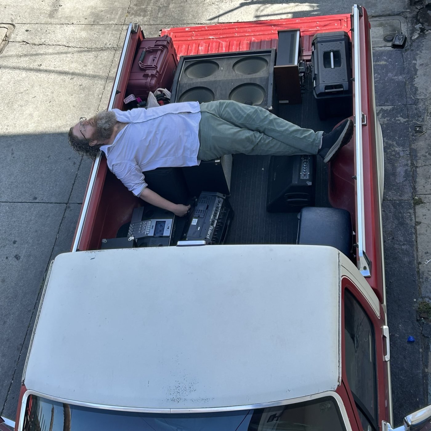 Overhead view of Aaron lying back in the bed of a red pickup truck among speakers, a road case and tape machines