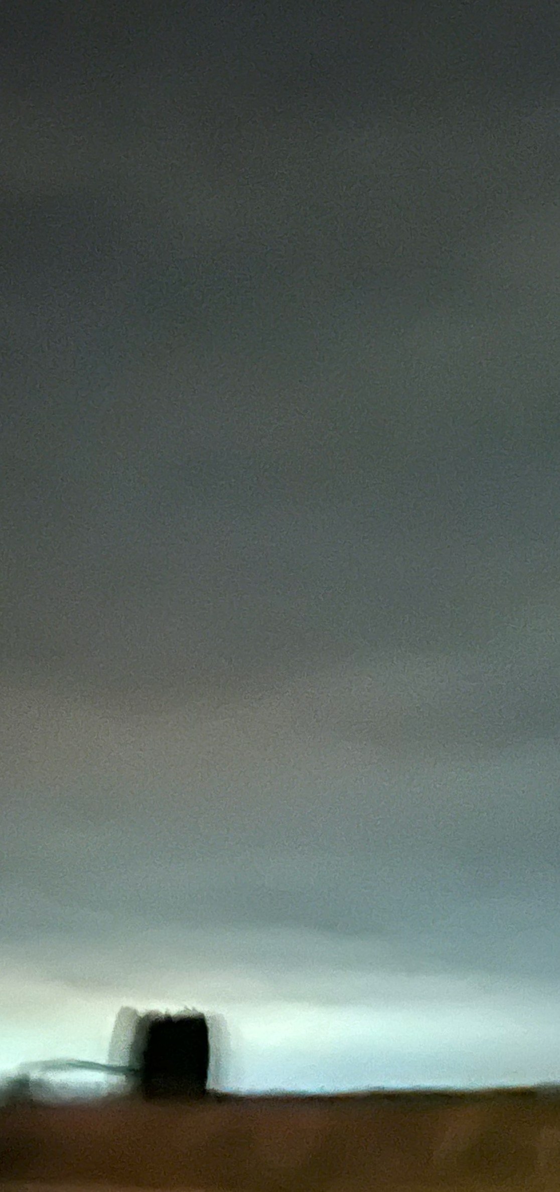 A blurred dark shape on a low horizon under a wide grey-green sky that brightens to a pale glow near the ground, with a warm brown band below