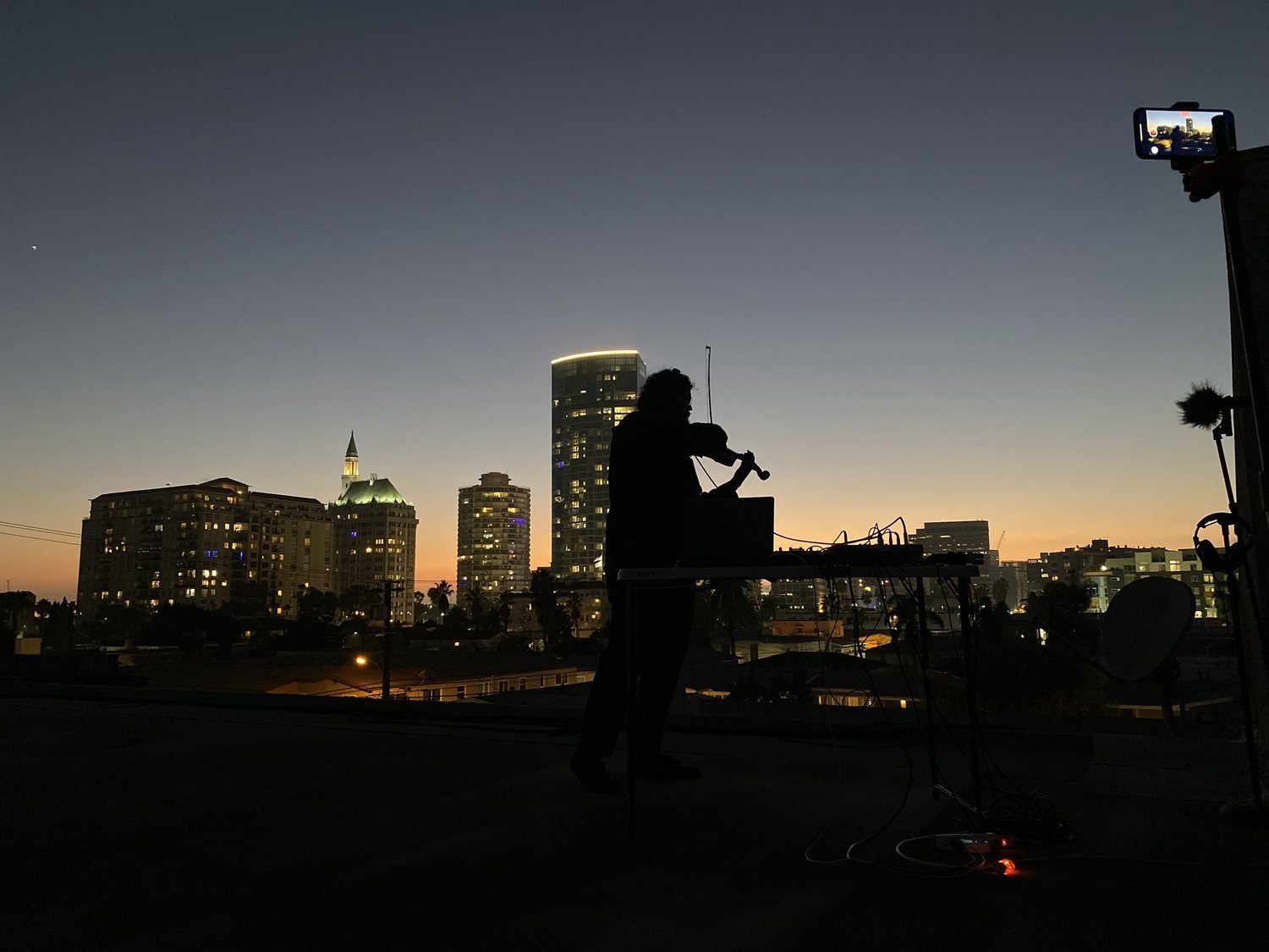 Silhouette of a violinist at a table of gear on a rooftop at dusk, city towers and an orange horizon behind