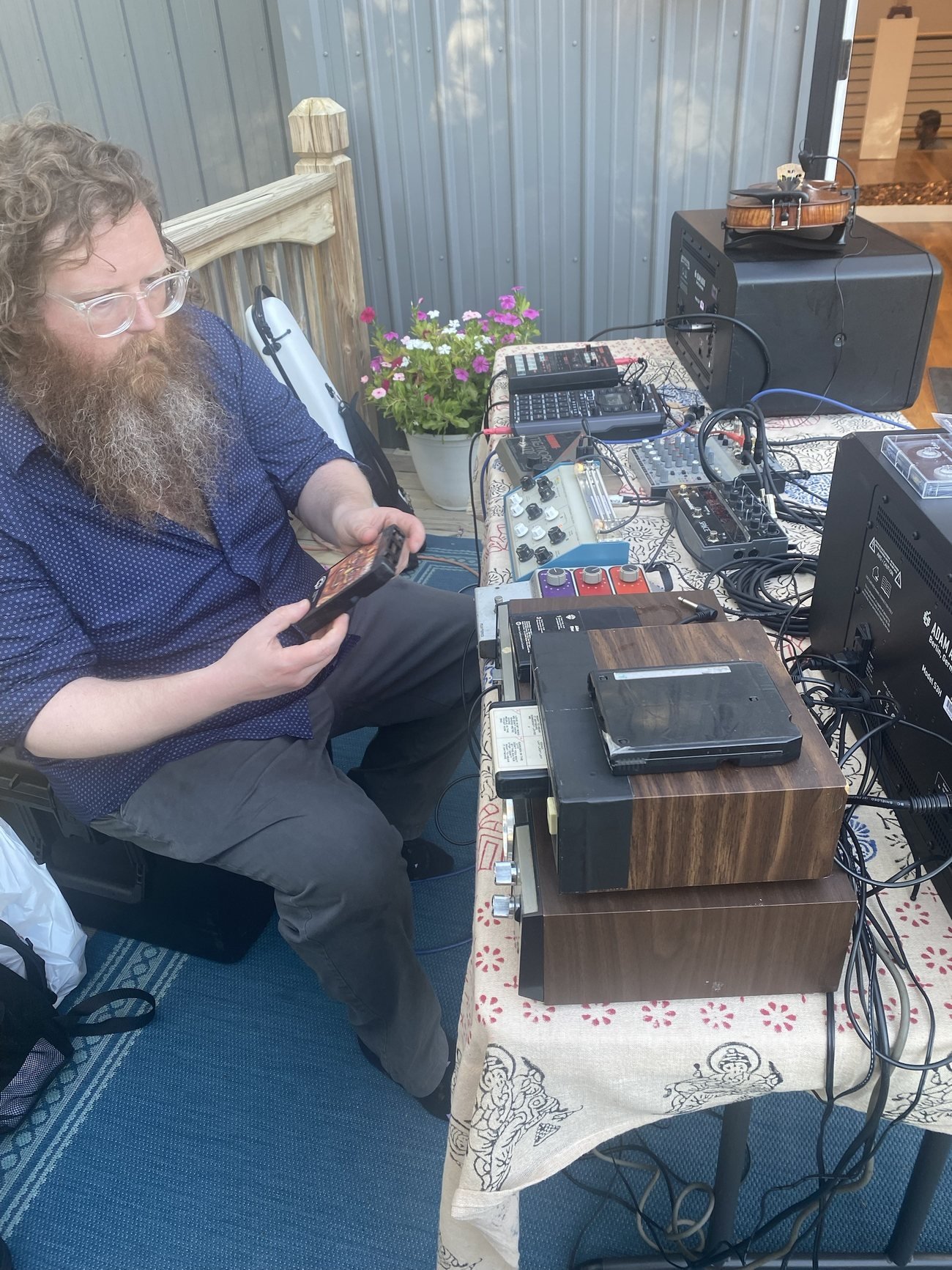 Aaron holding an 8-track cartridge while seated beside a table of pedals, a sampler and stacked 8-track players, his violin resting on a speaker behind