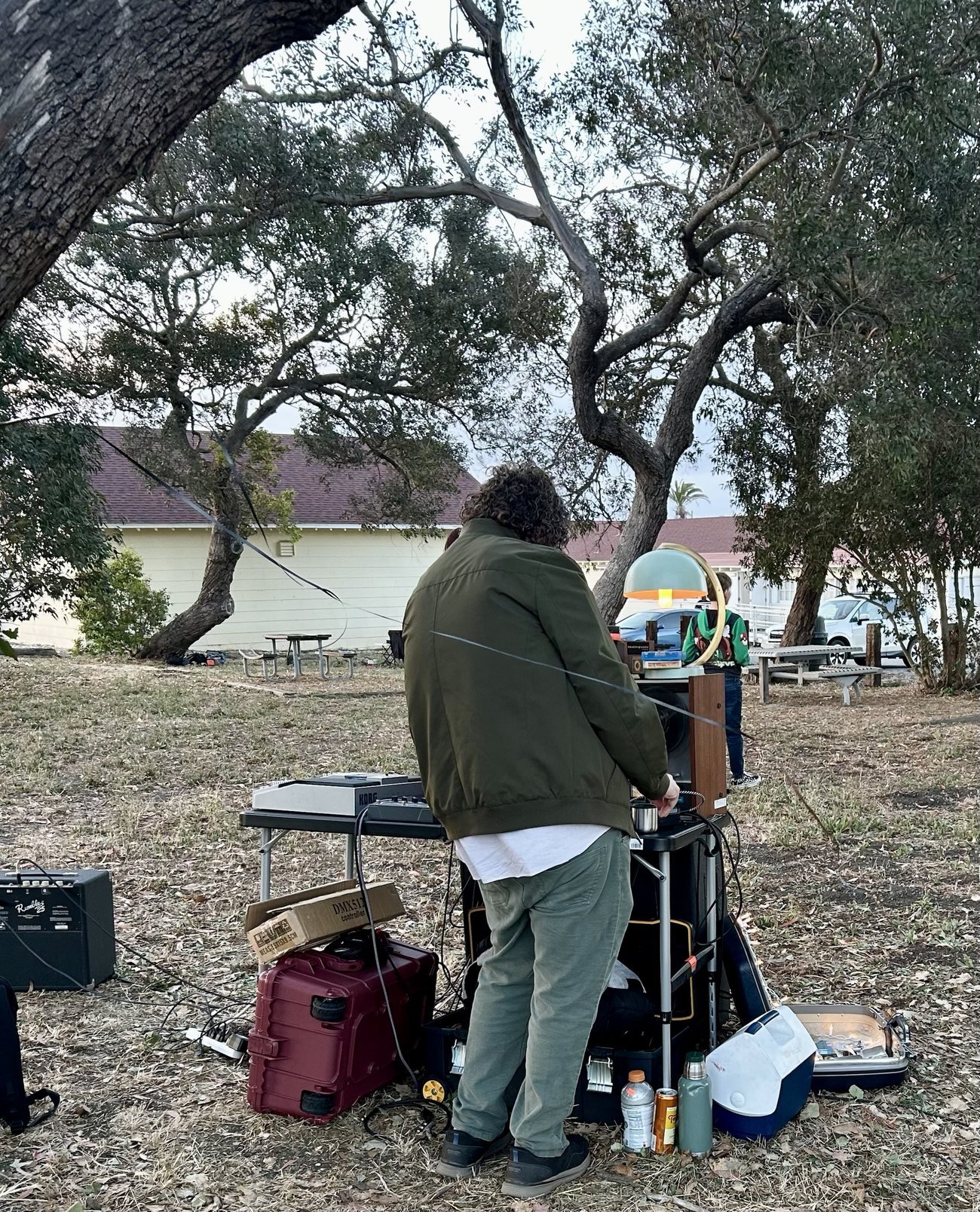 Seen from behind, Aaron adjusts equipment at a folding table under oak trees at dusk, with road cases, a cooler and a small amp on the ground
